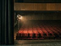 Leerer Zuschauerraum eines Theaters von der Bühne aus, rote Sitzreihen, eine einzelne Arbeitsleuchte